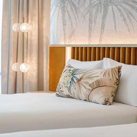 Amora Hotel Brisbane Zewnętrze zdjęcie The photo shows a stylish hotel room featuring two neatly made beds with white linens. Each bed has several pillows, one of which has a decorative cover with a tropical plant pattern. Behind the beds is a modern headboard, and the wall is decorated w