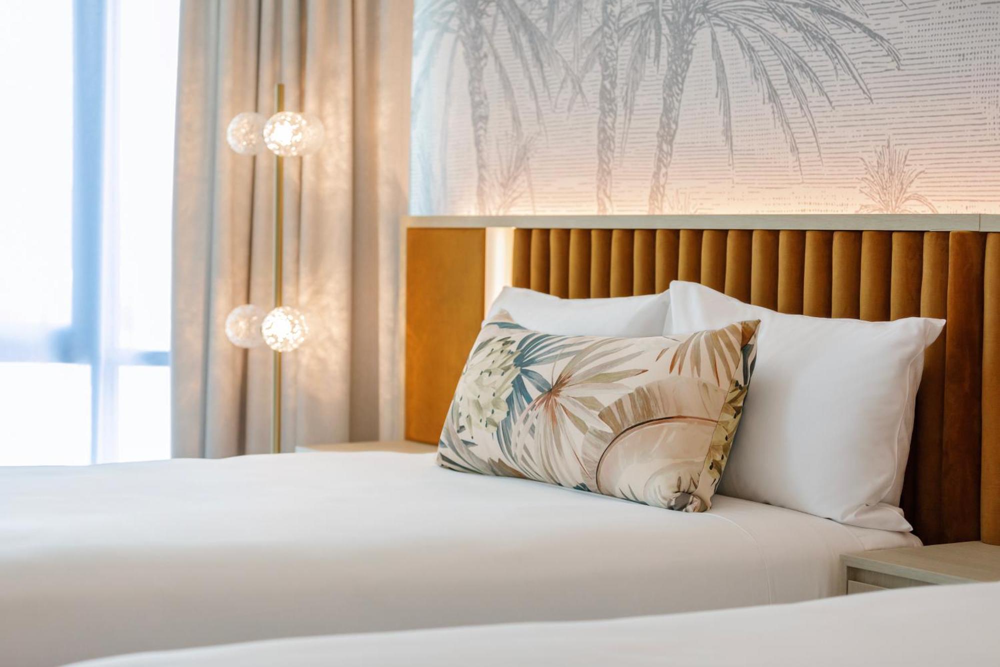 Amora Hotel Brisbane Zewnętrze zdjęcie The photo shows a stylish hotel room featuring two neatly made beds with white linens. Each bed has several pillows, one of which has a decorative cover with a tropical plant pattern. Behind the beds is a modern headboard, and the wall is decorated w