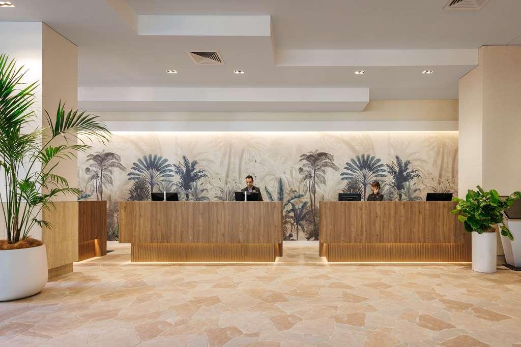 Amora Hotel Brisbane Wnętrze zdjęcie The photo shows a modern reception area with two wooden reception desks. Behind the desks, there is a decorative wall featuring a large mural of tropical plants and palm trees, creating a serene atmosphere. The space is well-lit with overhead lights,