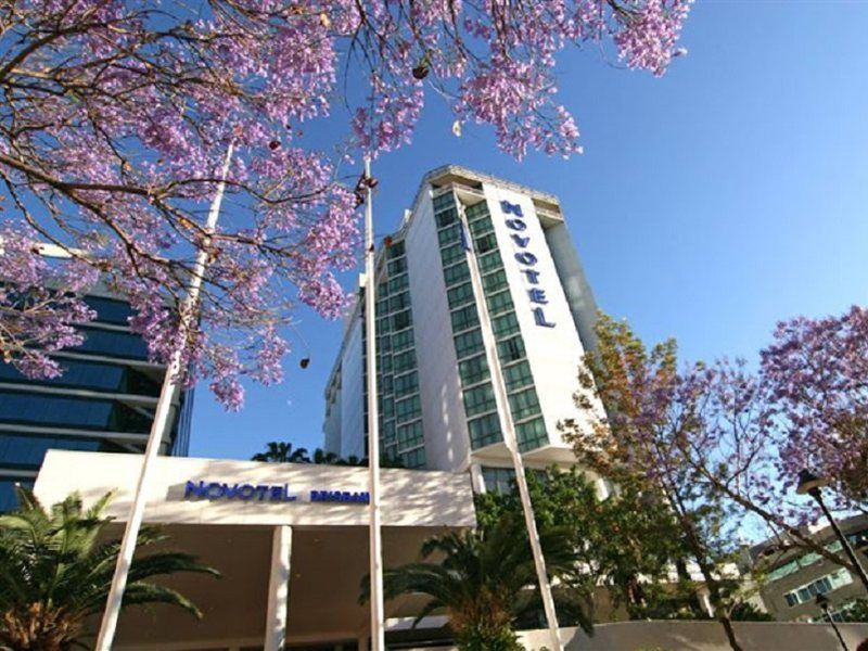 Amora Hotel Brisbane Zewnętrze zdjęcie The photo shows a modern hotel building, identified as "Novotel," rising above a landscaped area with palm trees. In the foreground, there are vibrant purple flowering trees, likely jacarandas, which add to the picturesque scene. The sky is clear and